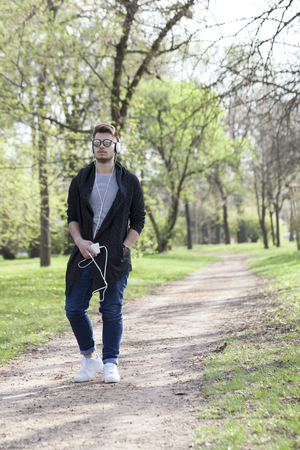 Young man relaxes by walking in the park, holding the phone in his hand and listen to music. Selective focus and small depth of field.の写真素材