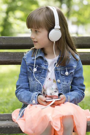 Little cute little girl with a nice face sits on the bench, listens to music and enjoys a beautiful day. She holds a mobile phone in her handの写真素材