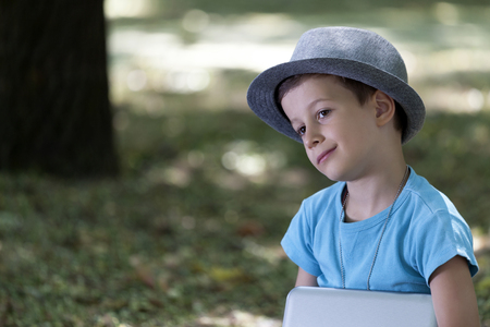 Sweet little  boy with hat sitting on the grass at sunset beautiful light in the park. Children's Day. He holds the tablet in his hand. Copy spaceの写真素材