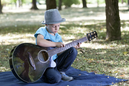 Portrait of a little sweet boy with hat sitting in a park, playing a guitar and enjoying a beautiful summer dayの写真素材