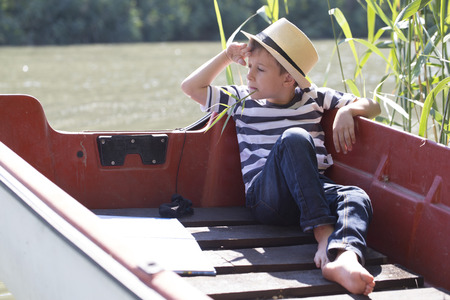 Little cute boy with hat sits in a boat on the river, holds the grass in his mouth and enjoys a beautiful and sunny dayの写真素材
