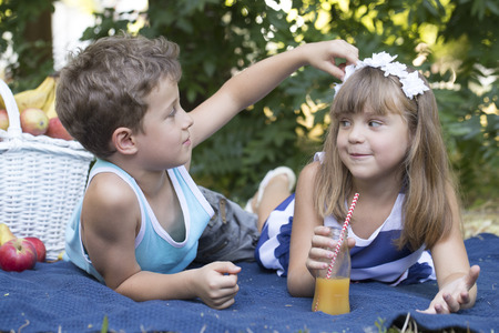 little cute boy and girl sit on a blanket in the grass and play.The boy looks at the small flowers in the girls hair . Near the boy and girl there is a picnic basket with juice and lots of fruit.First love, growing up, childhood, playing, socializingの写真素材
