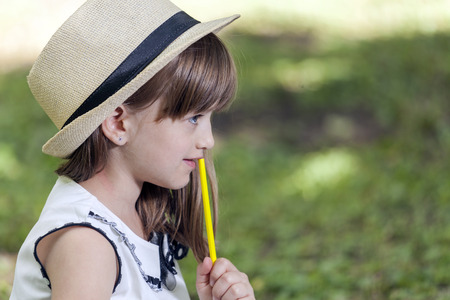 Close up portrait of a cute little girl with hat  in the park. She holds a wooden crayon, a pencil in her hand and looks thoughtfully. Copy spaceの写真素材
