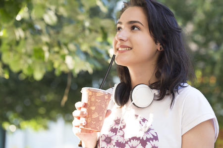Portrait of a beautiful young teenage girl with headphones around her neck outdoors. She was holding a large plastic cup with juice. With a beautiful smile and joyful emotions. Copy spaceの写真素材