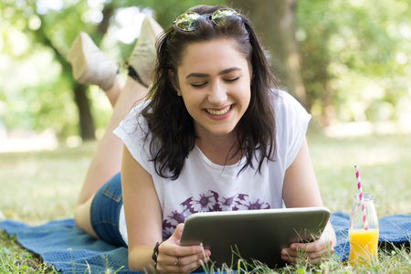 Beautiful young smiling girl - a teenager lying on a blanket in nature and using a tablet. hipster...