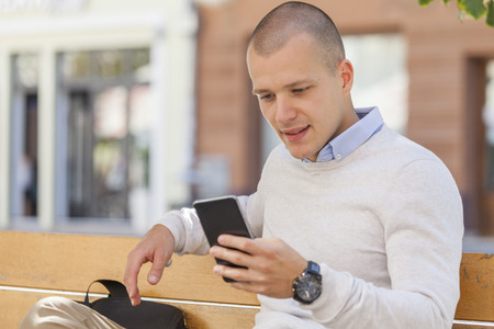 Portrait of an urban handsome young man sitting on the bench in the city and using a mobile phoneの写真素材