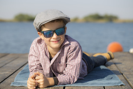 Portrait of a cute little boy with a hat and sunglasses, sitting by the river - the lake and enjoy the beautiful and sunny dayの写真素材