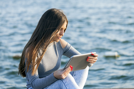 Young woman - teenager sitting by the ocean / river and use tablet. Woman use touch pad tablet pc internet technology. Copy spaceの写真素材