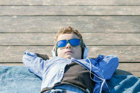 Little boy with sunglasses lying by the sea and enjoying a sunny day. He has headphones on his ears and listens to music.の写真素材