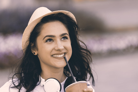 Close-up portrait of young beautiful teenage girl with hat and headphones. She is smiling and positive. In his hand, he holds a plastic cup from which he drinks a refreshing drink. Copy spaceの写真素材