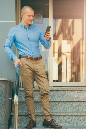 Young handsome businessman standing in front of the building and holding a mobile phone. He goes home from work and uses a cell phone to read new messages. Texting messageの写真素材