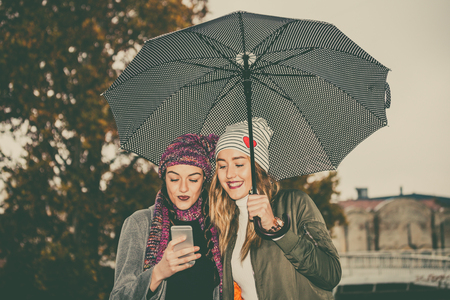 Two young women with umbrella and using mobile phone in the cityの写真素材