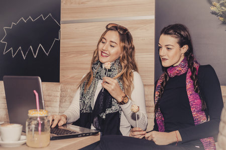 Two happy girls with cakes using laptop and drinking coffee in cafe. Friendship and technology