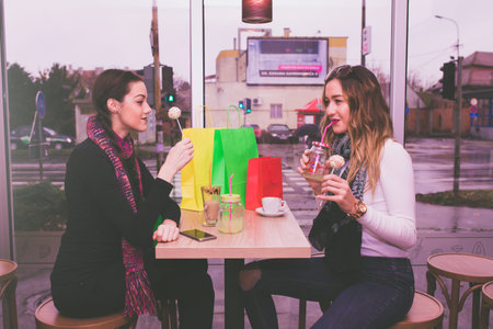 Two young happy girls with cakes and lemonade enjoying a conversation in cafe, after shopping
