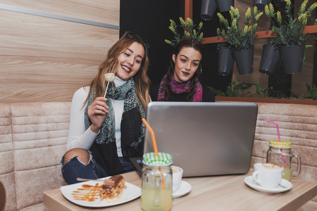 Two happy girls with cakes using laptop and drinking lemonade in cafe. Friendship and technologyの写真素材