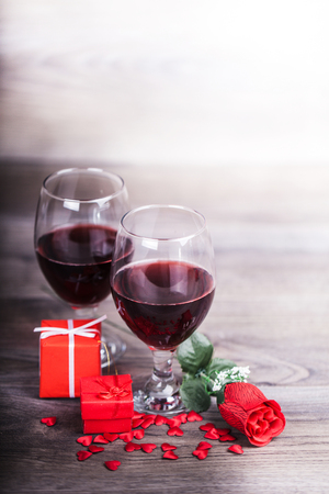 Two glasses of red wine, roseand gift with decoration by red hearts on wooden table. Valentines day celebration concept. Copy space.の写真素材