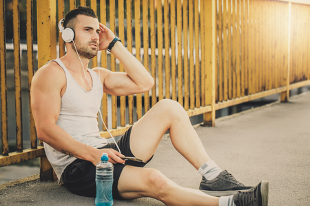 Fitness training. Young sportsman take a break after training. He siting on the bridge and listening to music with headphones. Fitness, sport, lifestyle conceptの写真素材