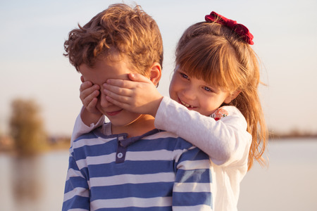 Girl and boy playing on the beach at sunset time. First love. Happy children enjoy while spending time near the lake and having a good time. Girl puts hands on boys eyes.の写真素材