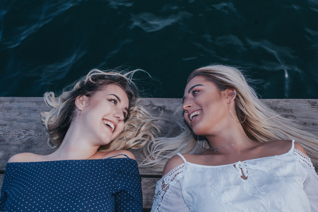 Friends having fun outdoors. Two beautiful girls lying next to the river and looking at each other. Laughing while enjoying the evening by the water. Lifestyle, vacation conceptの写真素材