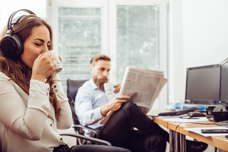 Businesswoman with headphones sitting at desk during the break and using mobile phone while man is reading newspaperの写真素材