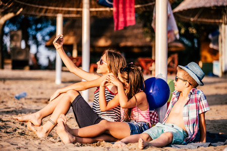 Portrait of positive children playing with mobile phones and making selfie on sandy beach togetherの写真素材