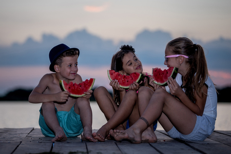 Happy children eating watermelon while sitting on a wooden pier and talking at sunset timeの写真素材