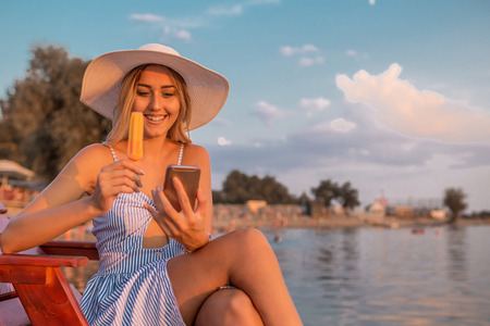 Attractive young woman in dress sitting on the wooden pier, using mobile phone and eating ice creamの写真素材