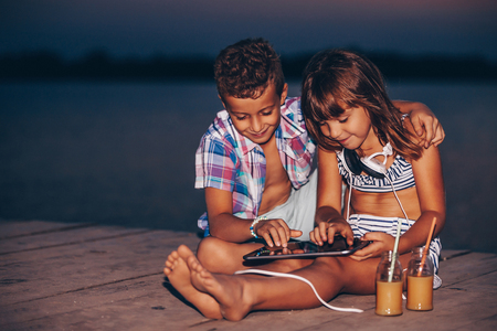Portrait of positive children playing with digital tablet together on wooden pier. Kids laughing, enjoying the beautiful sunsetの写真素材