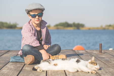Little cute boy is sitting by the river with his cat. They enjoy together on a beautiful sunny day. Growing up, love for animals - cats, free time, travel, vacation. Copy spaceの写真素材