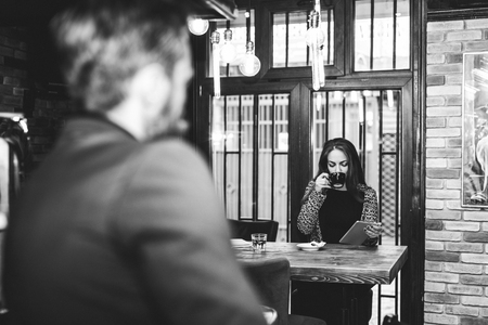 Young businesswoman sitting at the table in a cafe, using a digital tablet and drinking coffee, while a young man sitting at a bar and watching the beautiful womanの写真素材