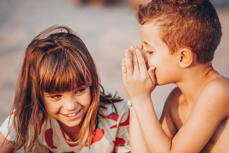 Little boy is whispering a secret into girl ear while playing on a sandy beachの写真素材