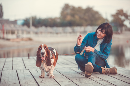 Portrait of beautiful young woman playing with dog by the river. Happy woman sitting on the wooden pier with her dog Basset Hound. Woman with puppy.の写真素材