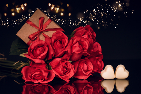 Valentines day greeting card with red roses, gift in a box and heart shaped chocolate  with lights on black backgroundの写真素材