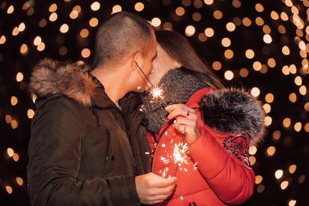 Young happy couple hugging and kissing. The young man and his girlfriend are having fun with sparklers in winter city night. - Imageの写真素材
