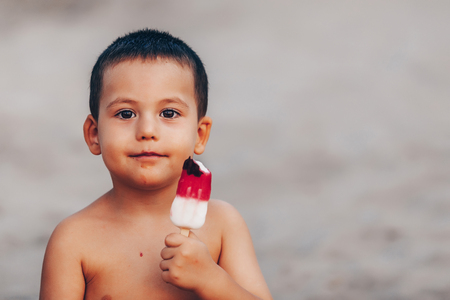 Cute Toddler boy Eating Ice-Cream. Kid with dirty face eating ice cream while sitting on sandy beach. Copy spaceの写真素材