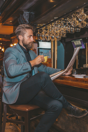 Portrait of a young bearded businessman sitting at the bar, reading newspapers and drinking coffee.の写真素材