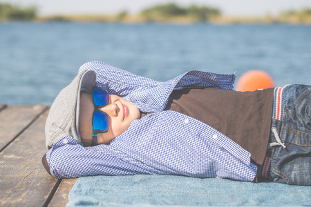 Portrait of a cute little boy with a cap and sunglases, lying by the river and enjoy the beautiful and sunny dayの写真素材