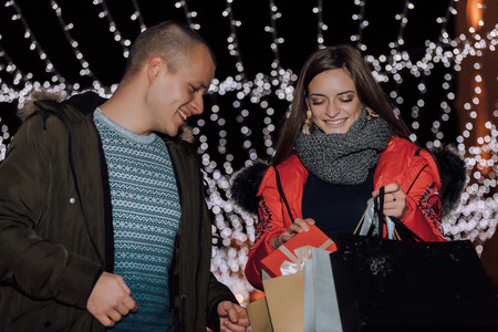 Young cheerful couple holding shopping bags, gift in a box and having fun at the night in the city street with a lot of lights on background. - Imageの写真素材