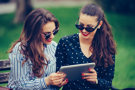 Two beautiful women laughing watching media content together in a digital tablet,  sitting on a bench in the park. - Imageの写真素材