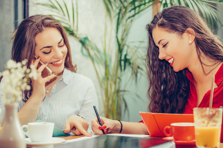 Happy female friends, students reading notes together, pointing where to sign a contract, talking and drinking coffee while sitting in the table in cafe. - Imageの写真素材