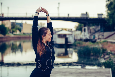 Young beautiful woman doing stretching her arms while listening to music by the river, preparing for...