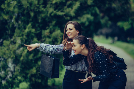Two female best friend walking through the park, after shopping. They smiling and pointing away in...