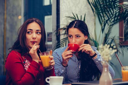 Image of a two happy girls friends sitting in cafe talking with each other drinking coffee,  looking at someone.の写真素材