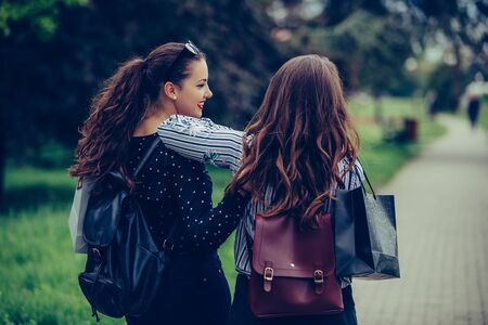 Back view of a two beautiful female friends hugging each other while walking through the park, after shopping.の写真素材
