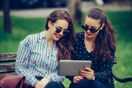 Two beautiful women laughing watching media content together in a digital tablet, sitting on a bench in the park.の写真素材
