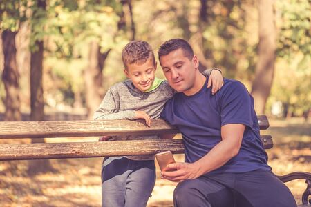 Little boy and his father taking a break after workout and watching something on smart phone in the park. Father and son spend time together and lead a healthy lifestyle.の写真素材