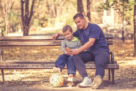 Little boy and his father taking a break after workout and watching something on smart phone in the park. Father and son spend time together and lead a healthy lifestyle.の写真素材