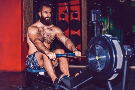 Bearded young man using rowing machine in the gymの写真素材
