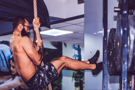 Young bearded man climbing on the rope during training in the sport gym