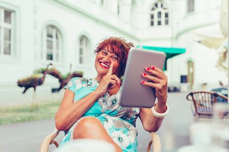Close up portrait of happy senior woman with finger to lips while making a video call with digital tablet in outdoor cafeの写真素材